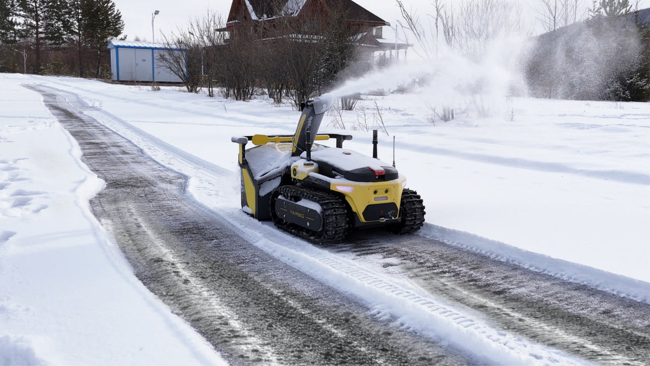 Yellow snow blower clears snow from gravel driveway, revealing clean path beside snowy yard.