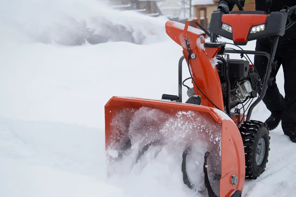 Bright orange self-propelled snow blower clearing deep snow, operator guiding it through winter storm.