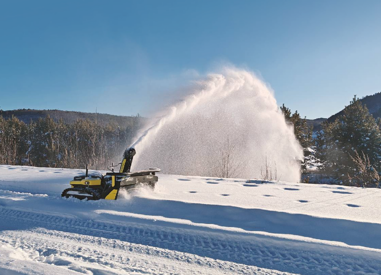 Yellow snowblower with steel chute powers through heavy wet snow, spraying plume high under clear blue sky.