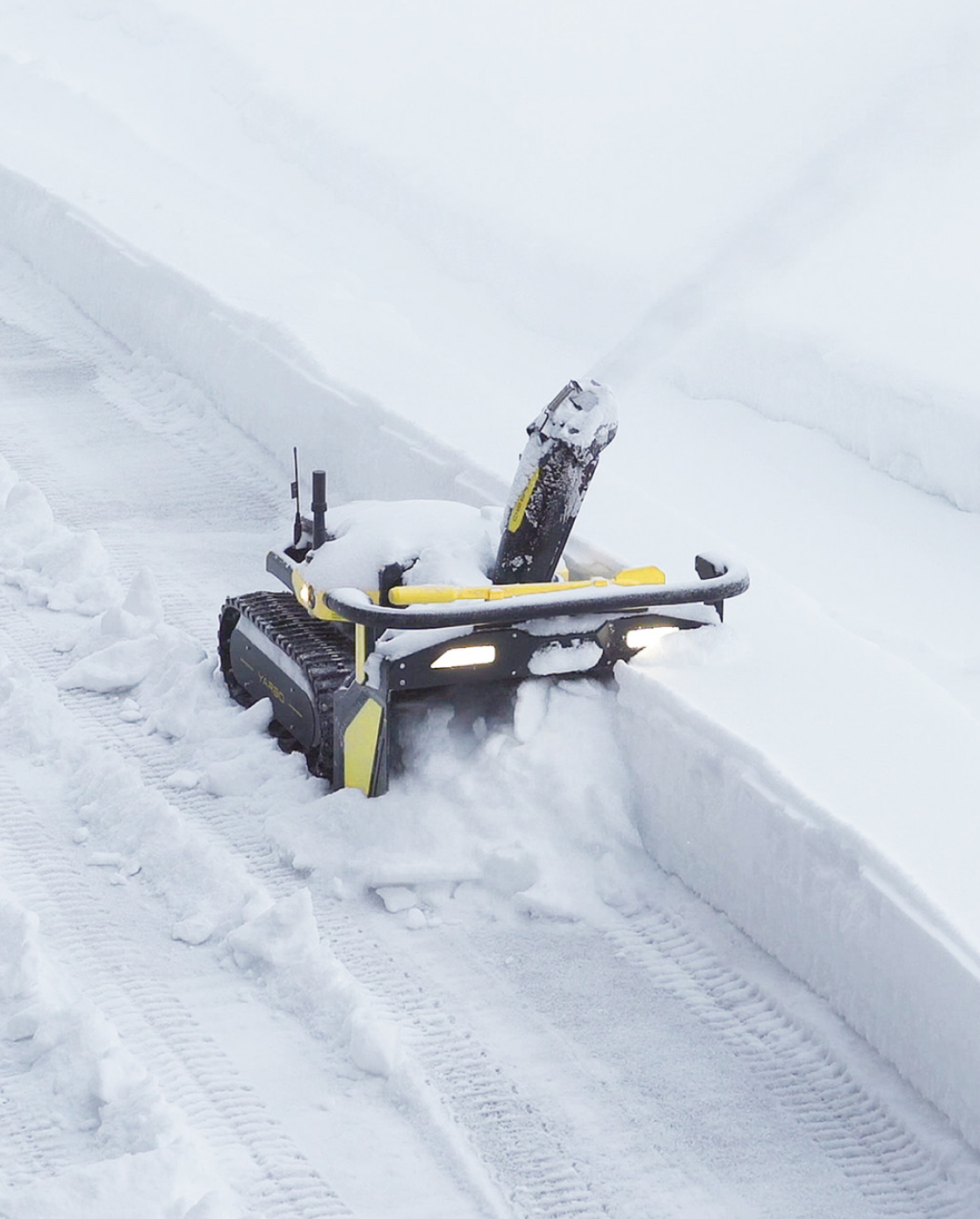 Black and yellow robotic snow blower by Yarbo clearing snow in a driveway, side view with illuminated headlights and tracked wheels.