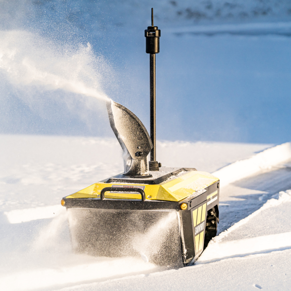 Yellow snow blower ejecting snow in bright sunlight on a snowy slope, with a tall black antenna on top.