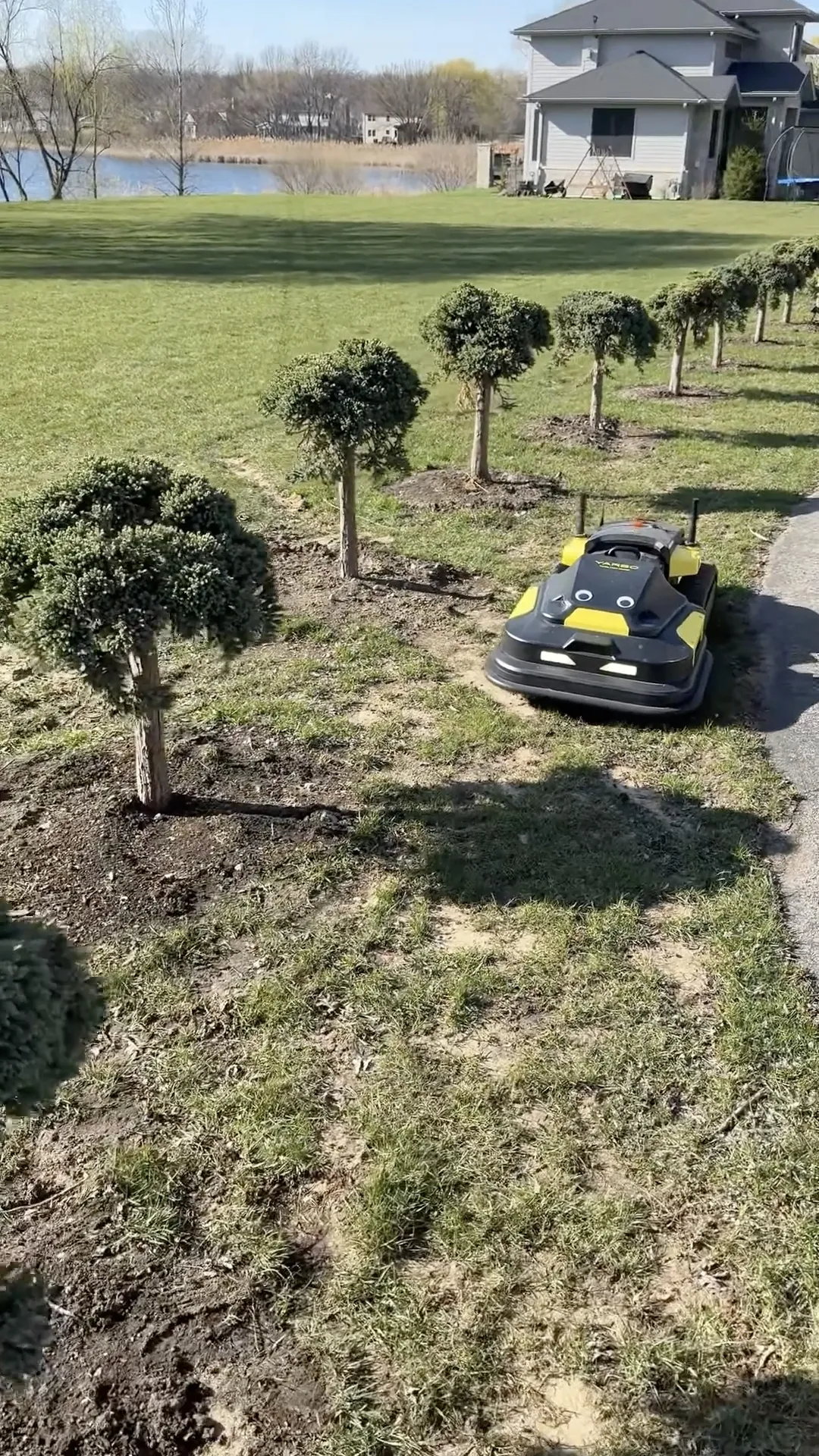 Black and yellow robotic lawn mower on grass near trimmed trees, house, and lake in background.