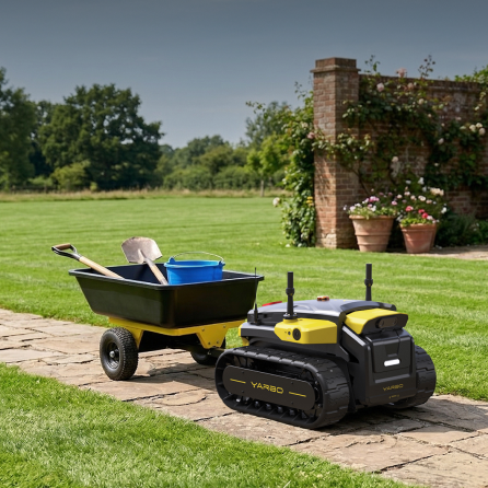 Black and yellow YARBO robotic lawn mower on stone path beside wheelbarrow with tools, green lawn and brick wall in background.
