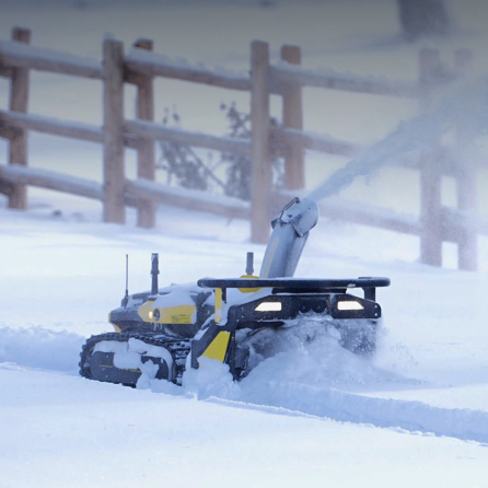 Yellow and black snow blower clearing snow in winter, tracks visible, wooden fence in background.