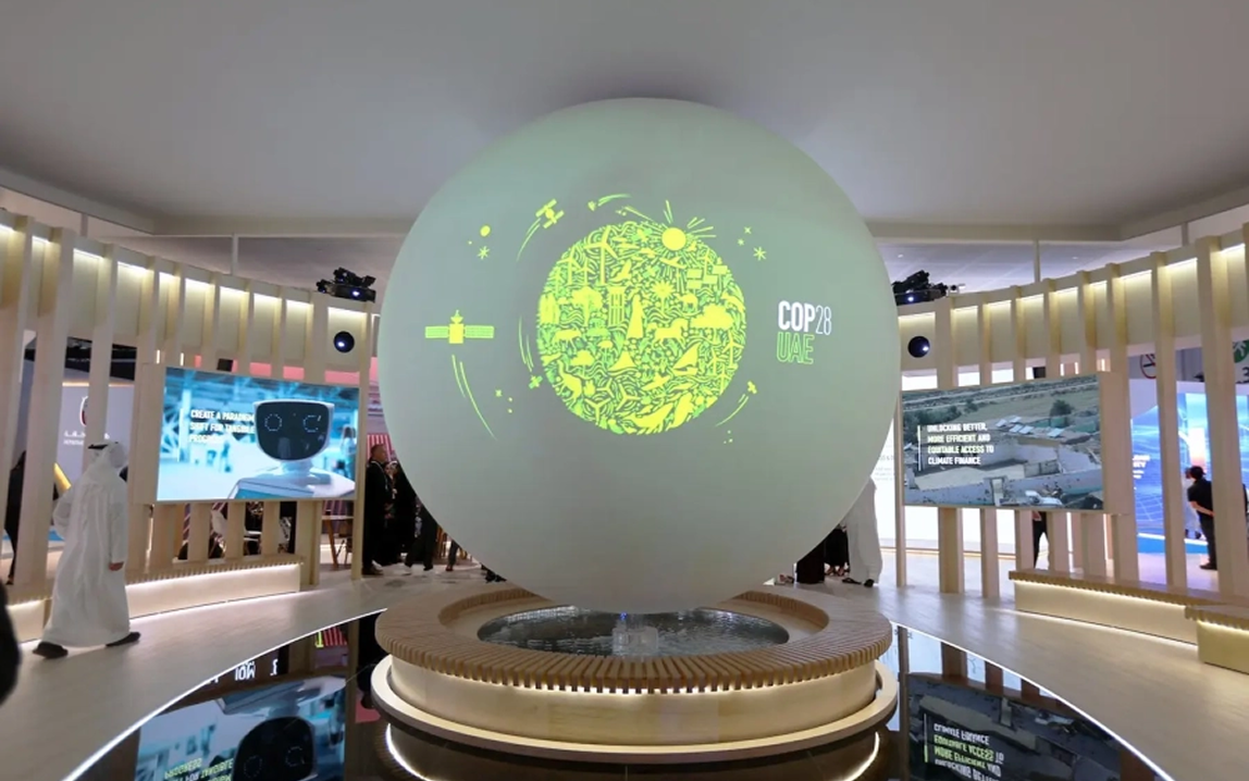 Large illuminated globe with green Earth design and "COP28 UAE" text, displayed in modern exhibition space with digital screens and visitors.