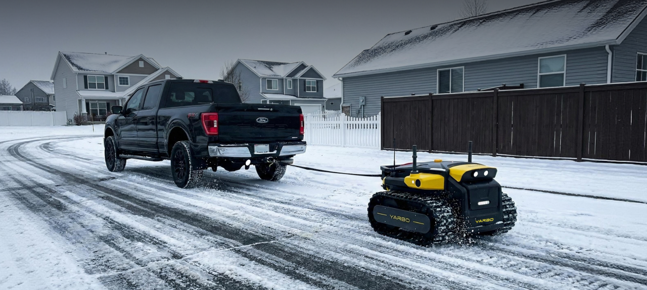 Black Ford truck towing yellow and black YARBO snow-clearing robot on snowy suburban driveway.