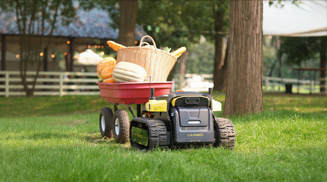 Black and yellow YARBO robot pulls red wagon with pumpkins and wicker basket on green lawn.