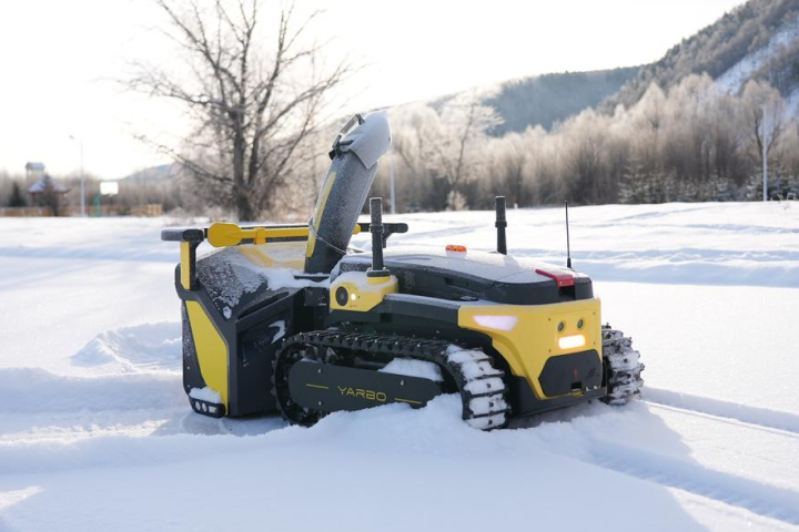 Yellow and black YARBO robotic snowplow clearing snow in a serene, frost-covered mountain landscape.