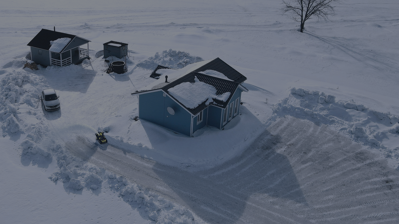 Aerial view of a snow-covered yard with a remote control snow plow robot clearing a path beside a blue house.