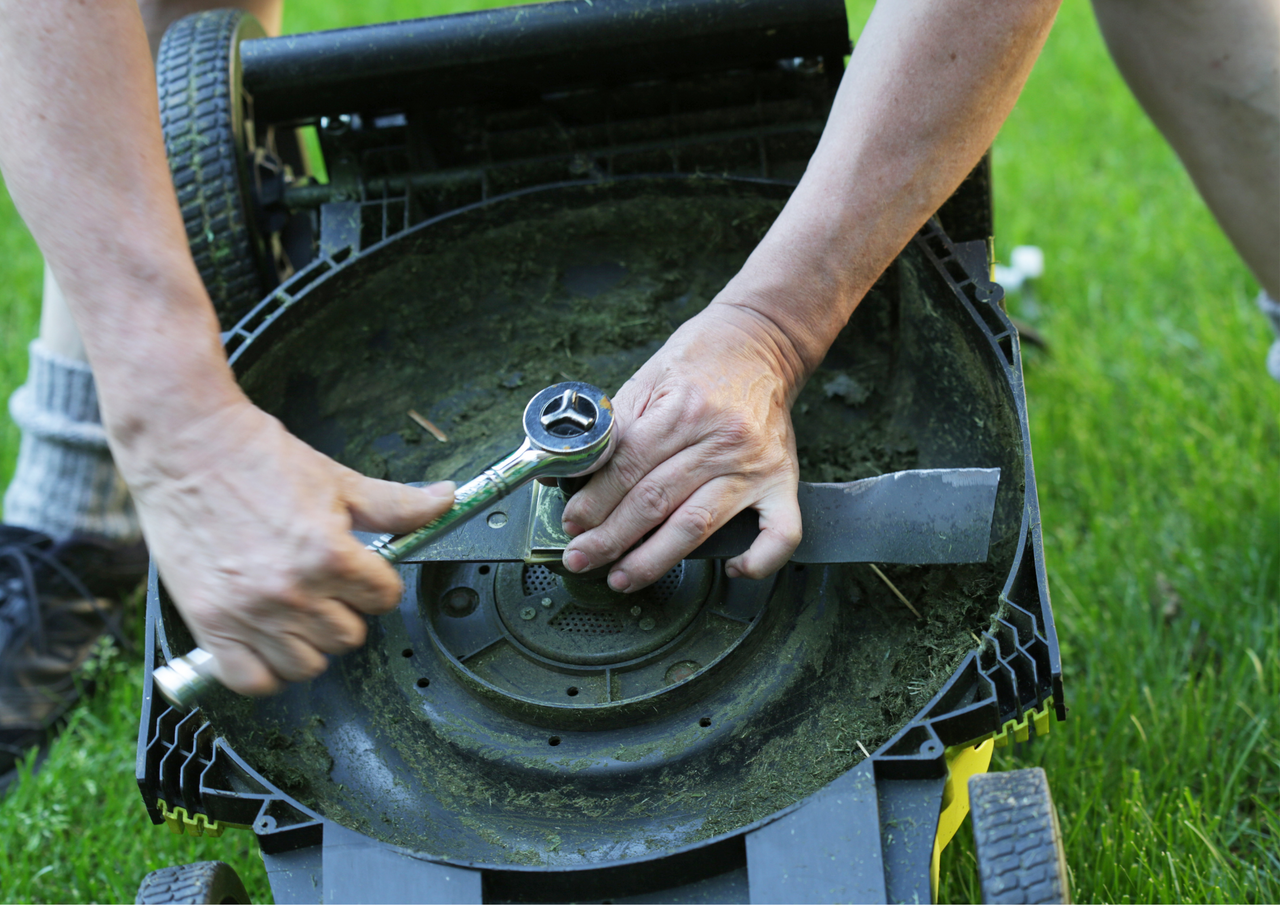 Hands tightening a mower blade with a socket wrench on a grassy lawn, ensuring a sharp, clean cut.
