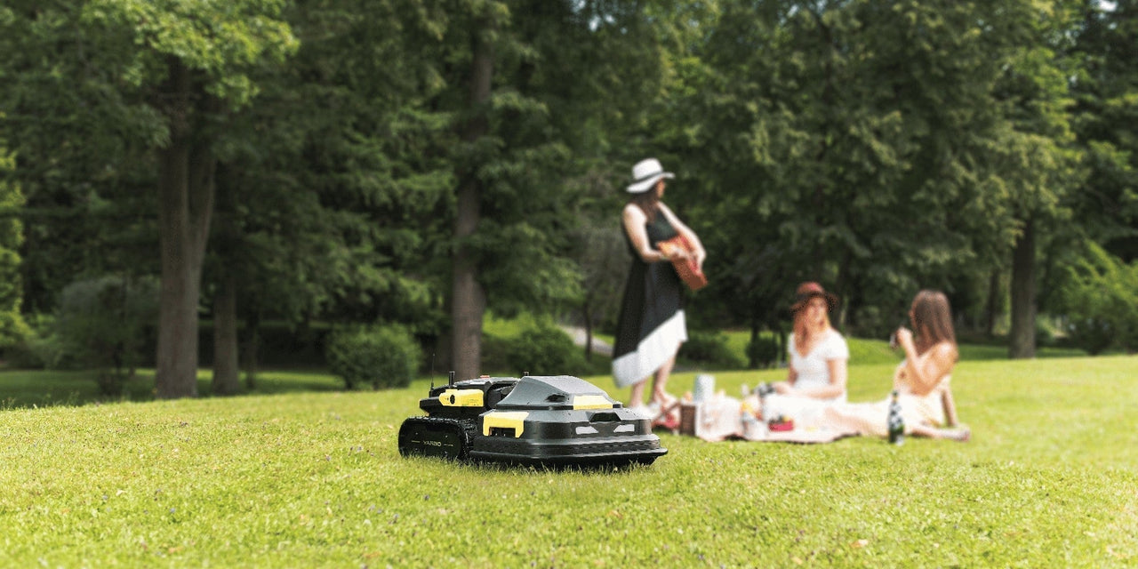 Robot lawn mower on vibrant green grass, with women enjoying a picnic in the background under leafy trees.