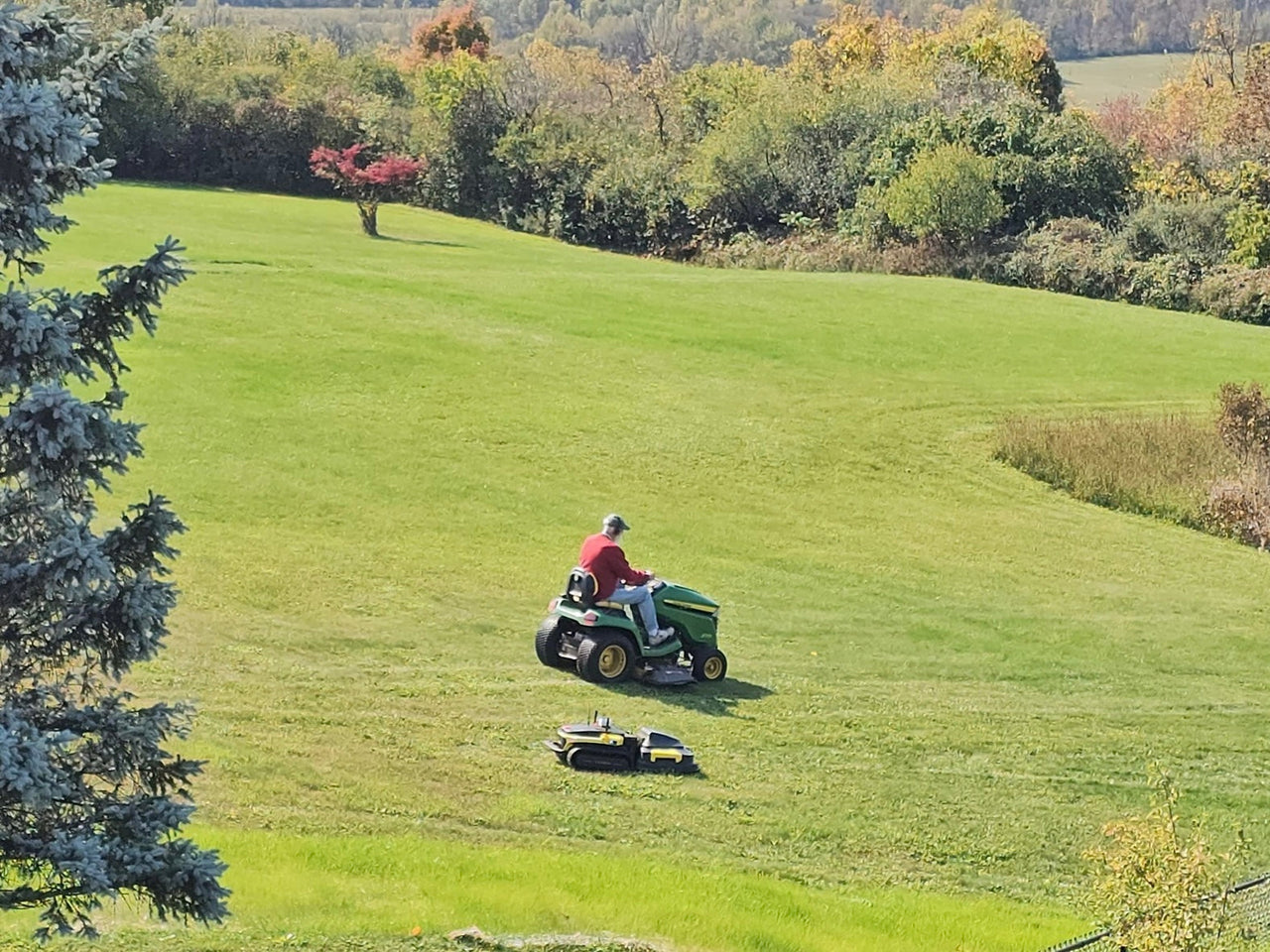 Man mowing vibrant green lawn with riding mower, robot mower nearby, autumn trees in background.