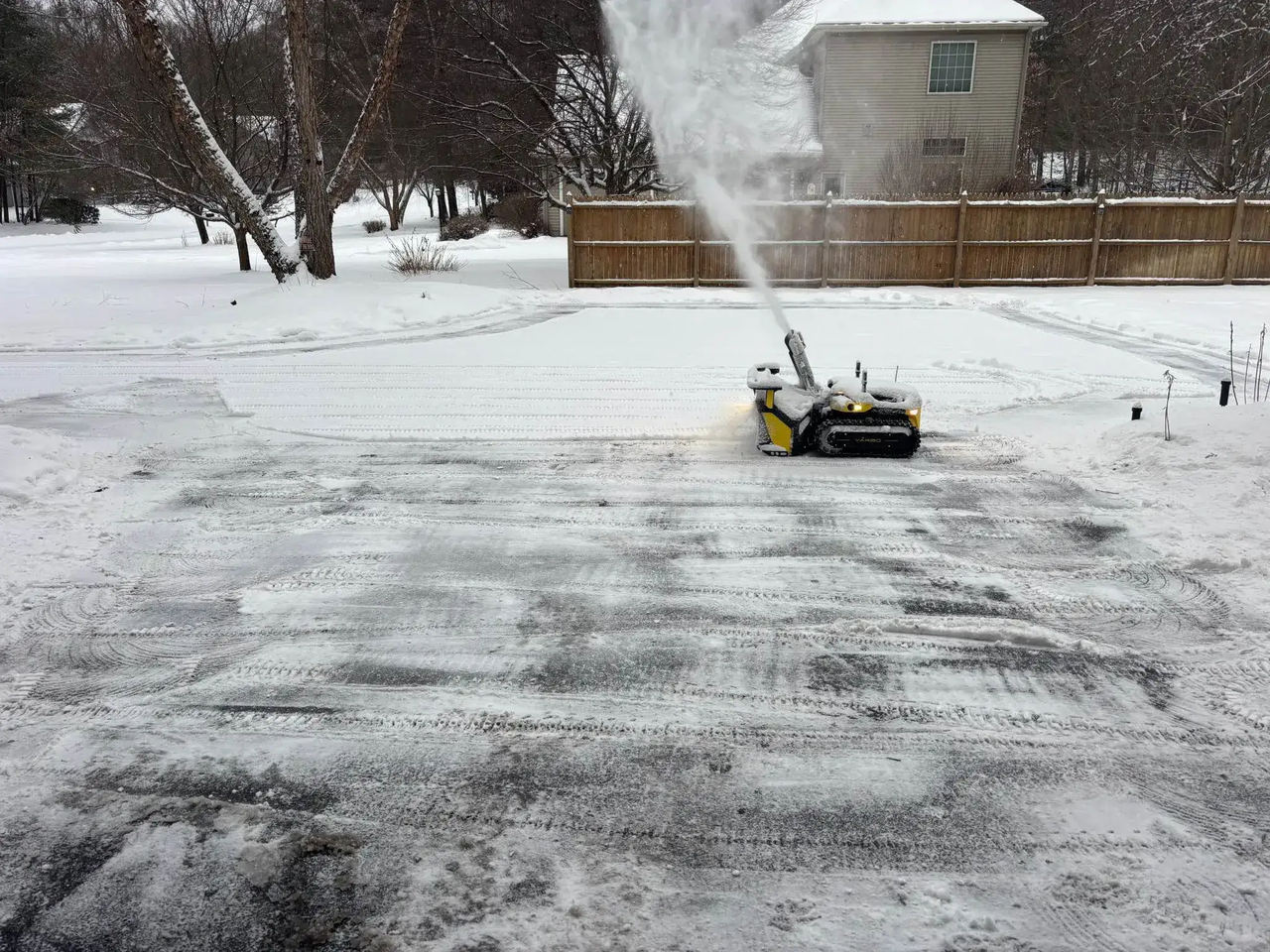 Yellow snow blower clearing driveway, spraying snow into air on snowy winter day.