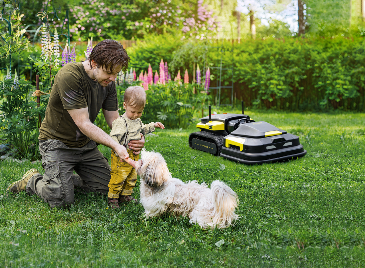 Man and child petting dog in lush garden, robotic lawn mowers nearby, sunny day.