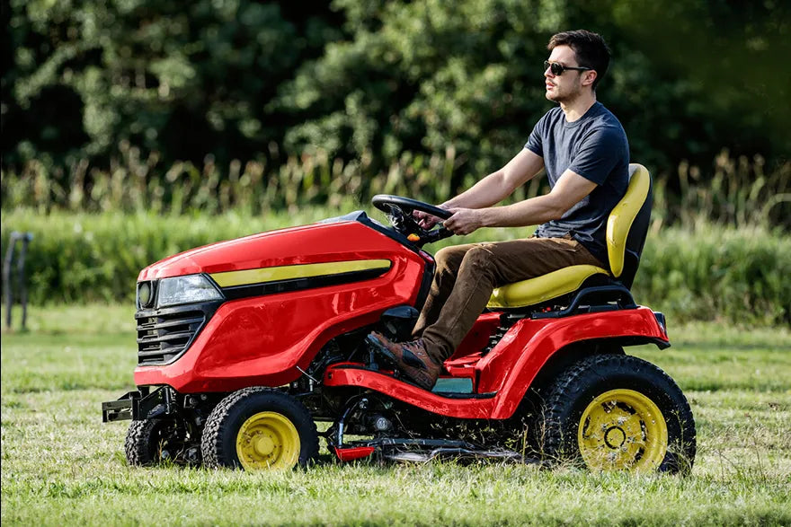 Man driving red riding lawn mower with yellow accents across lush green lawn on sunny day.