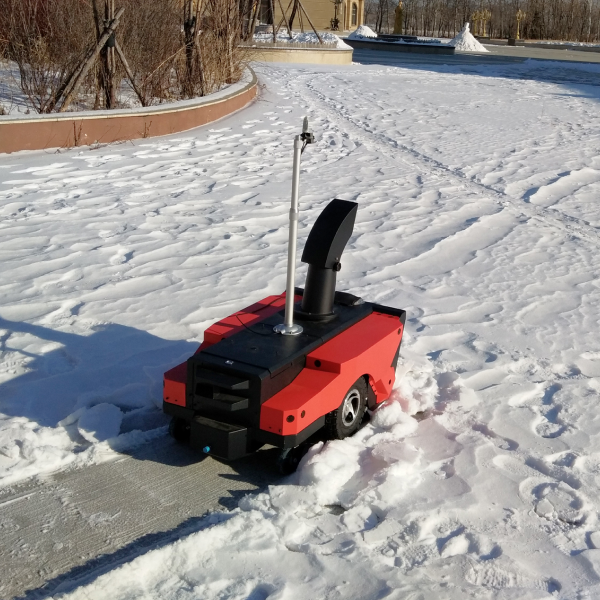 Red and black robotic snow blower clearing path in snowy park, bright sunlight, tracks visible.