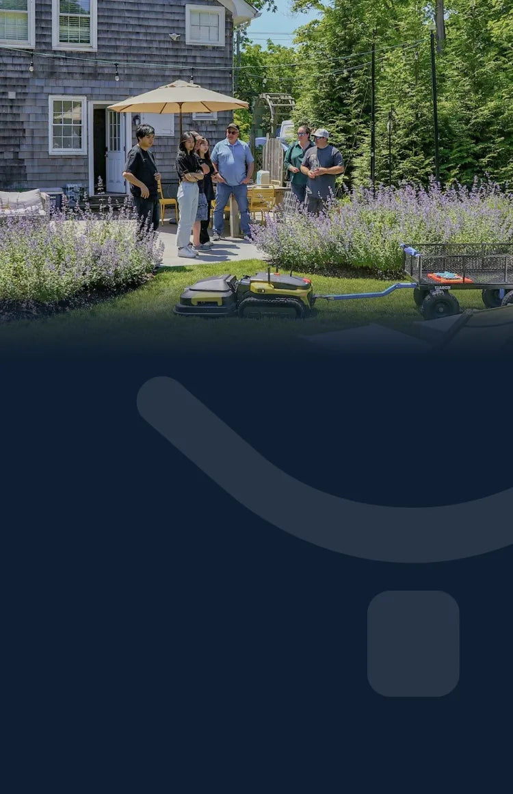 Group of people gather on a sunny patio beside a gray shingled house, with purple flowers, a yellow umbrella, and two robotic mowers in the foreground.