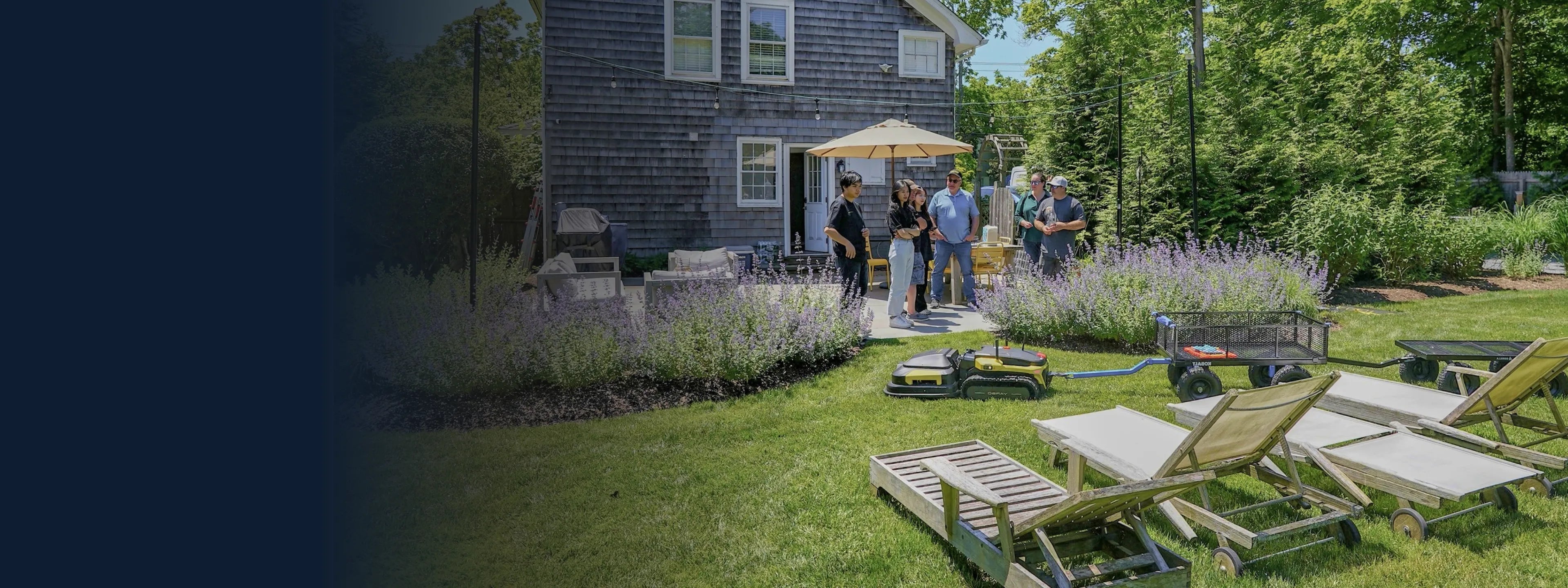 Group gathers on patio beside gray shingle house, surrounded by lavender, lawn chairs, and robotic mowers under sunny sky.