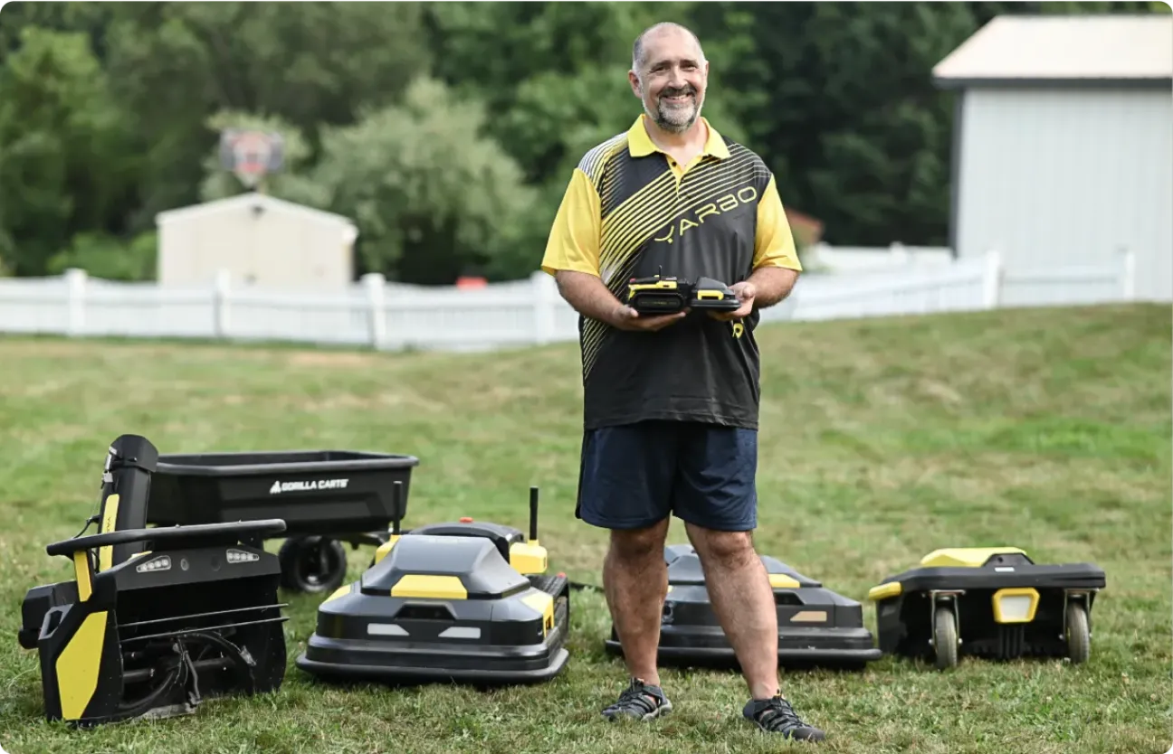 Man in yellow and black shirt stands on grass holding remote, surrounded by black and yellow robotic lawn mowers and a cart.