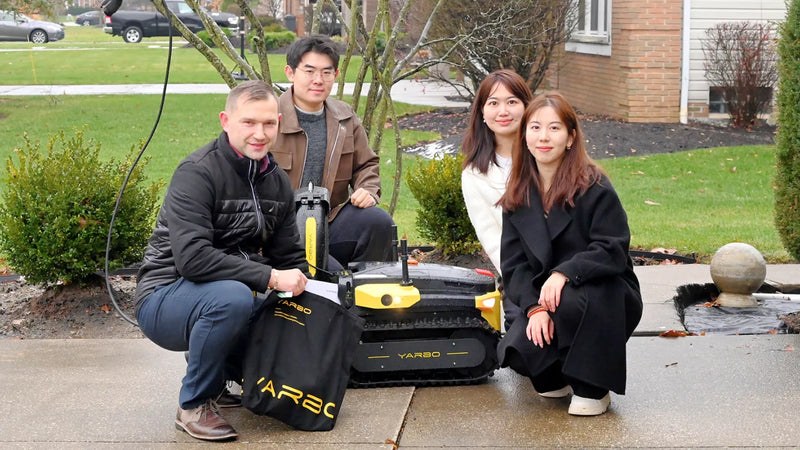 Four people pose with a black and yellow YARBO robot on a sidewalk, holding a branded bag, in front of a house and green lawn.