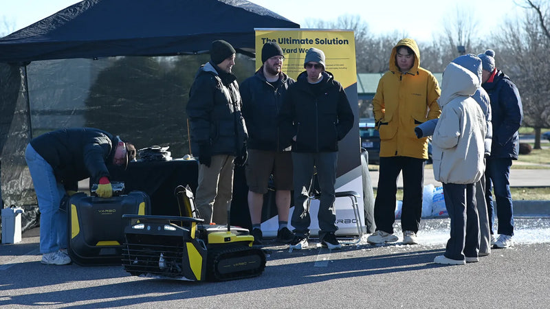 People gather at an outdoor booth under a tent, viewing a yellow and black YARBO robotic lawn mower on display.