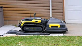 Black and yellow robotic lawn mower with tracks sits on concrete patio beside grass, near brown siding and white garage door.
