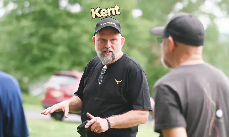 Man with gray beard and black cap labeled "Kent" gestures while speaking outdoors, wearing black shirt with gold logo, sunglasses on chest, green trees and red car in background.