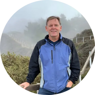 Man in blue and black jacket smiles outdoors on foggy hillside, hand on railing.