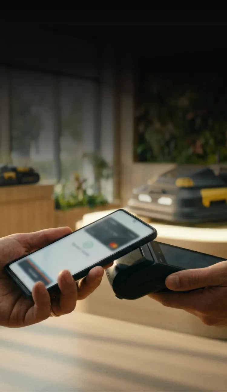Hands holding smartphone and payment terminal, completing contactless transaction with Mastercard logo visible, in warm-lit indoor setting.