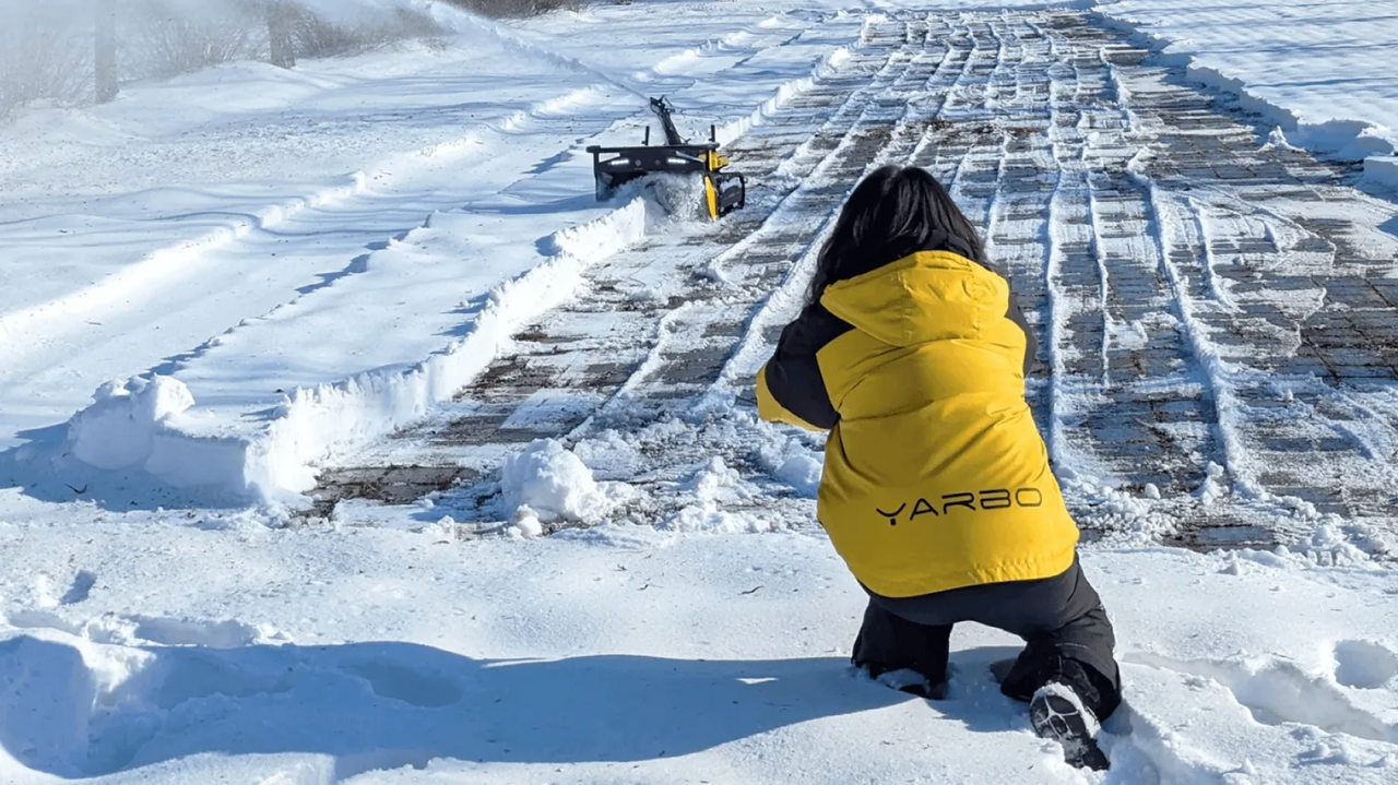 Person in yellow YARBO jacket kneels in snow, watching snowplow clear brick path. Bright sunlight, winter scene.
