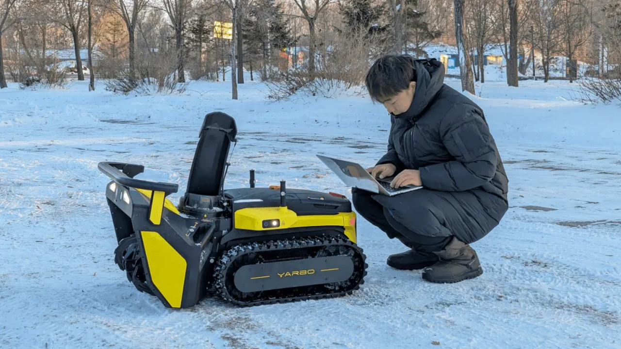 Man in black winter coat crouches in snow, using laptop beside yellow and black YARBO tracked robot.