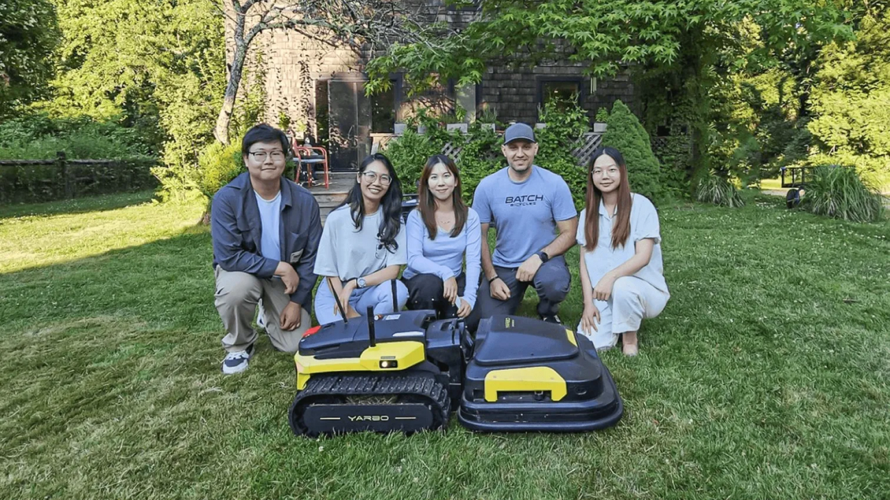 Five people crouch on a grassy lawn behind two black and yellow robotic lawnmowers, with a stone house and green trees in the background.