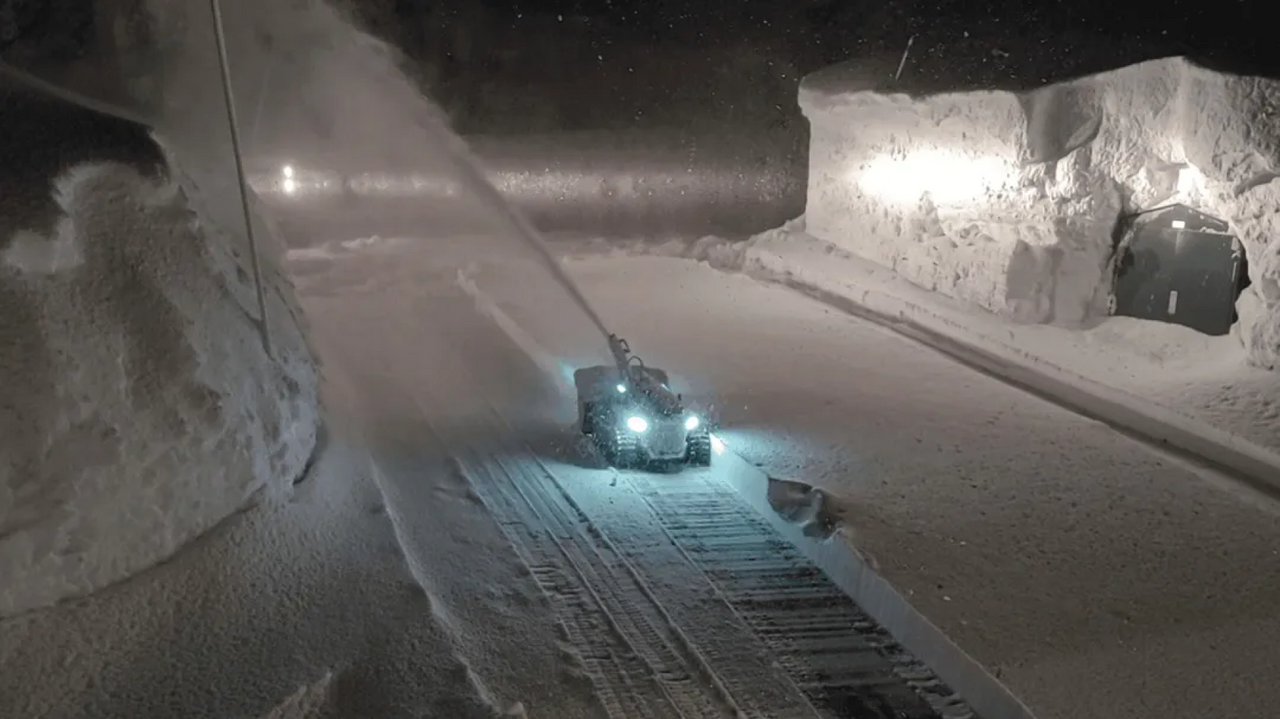 Snowplow clears icy road at night, headlights illuminating snowbanks and wet pavement.