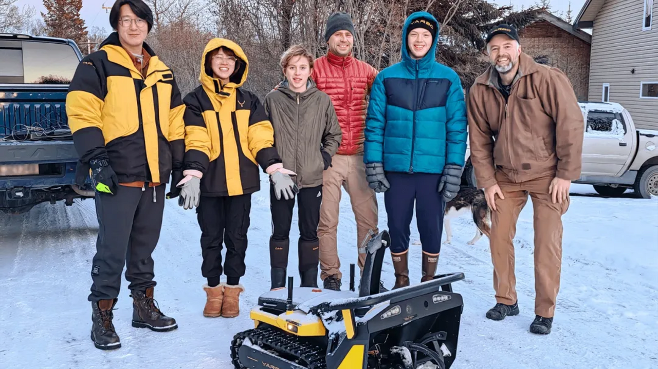 Six people in winter gear stand in snow beside a yellow and black tracked robot, with trucks and a house in the background.