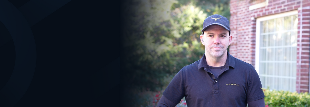 Man in black YARBO polo and cap stands outdoors near brick building with window, greenery in background.