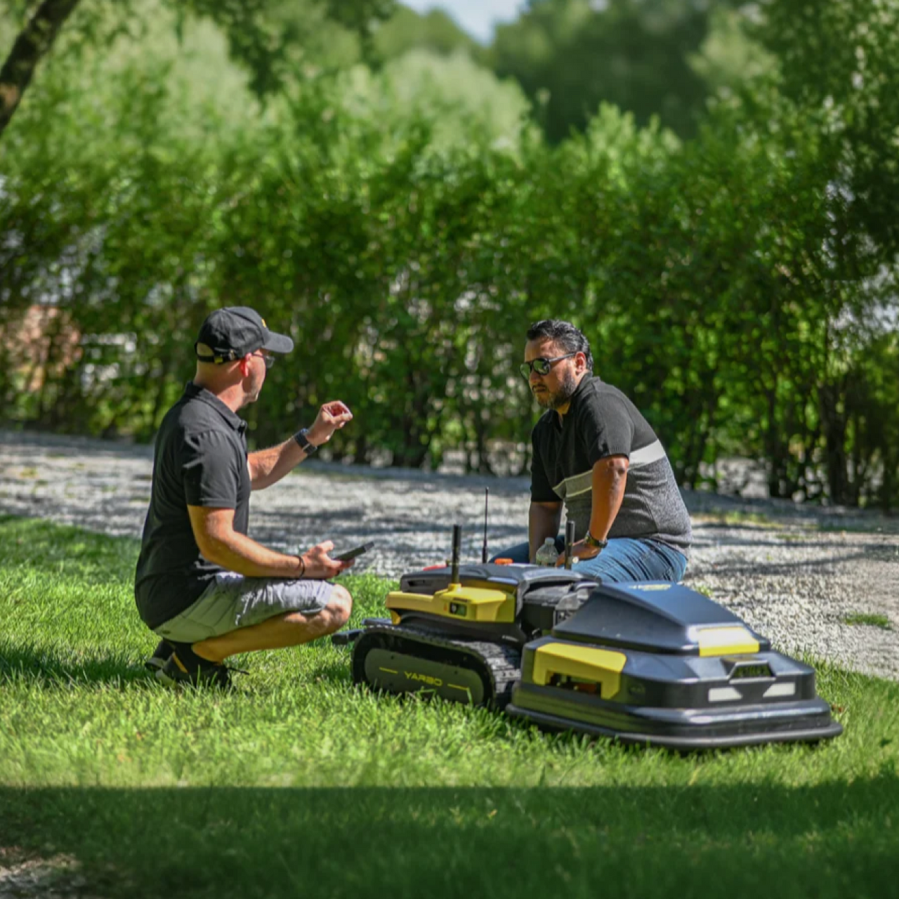 Two men crouch on grass, examining yellow and black robotic lawn mowers under bright sunlight.