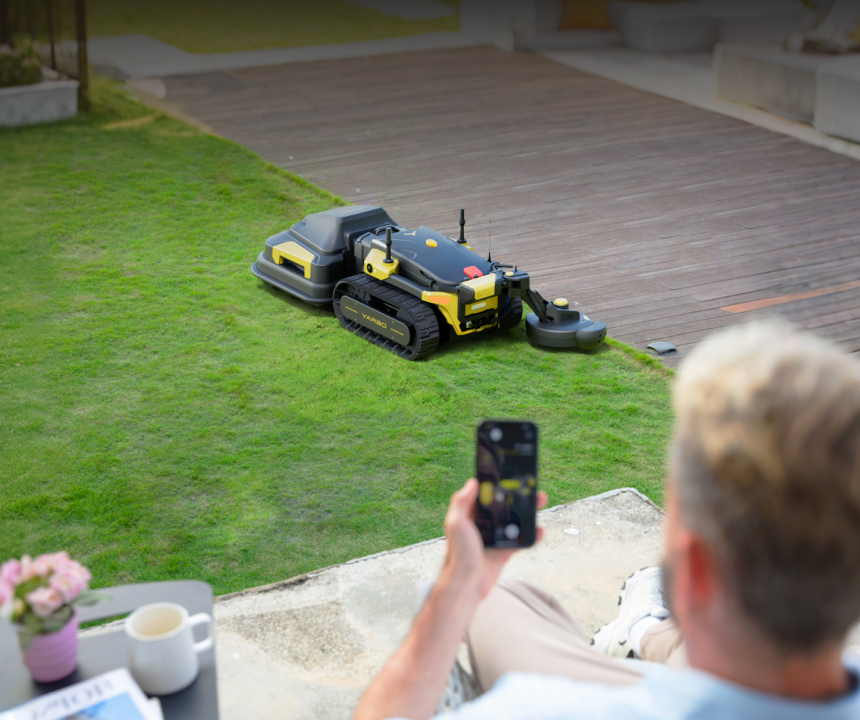Man relaxes on patio, watching black and yellow robotic lawn mower on green grass, using smartphone to control it.
