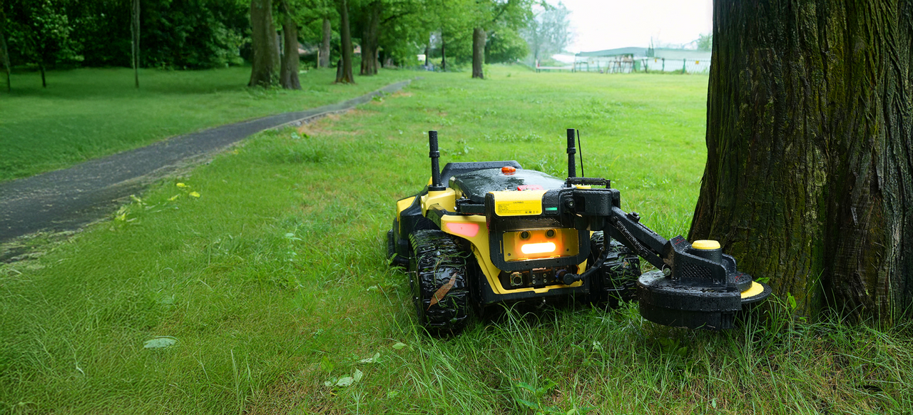 Yellow and black tracked robot with illuminated front light rests on grass beside tree trunk in park.