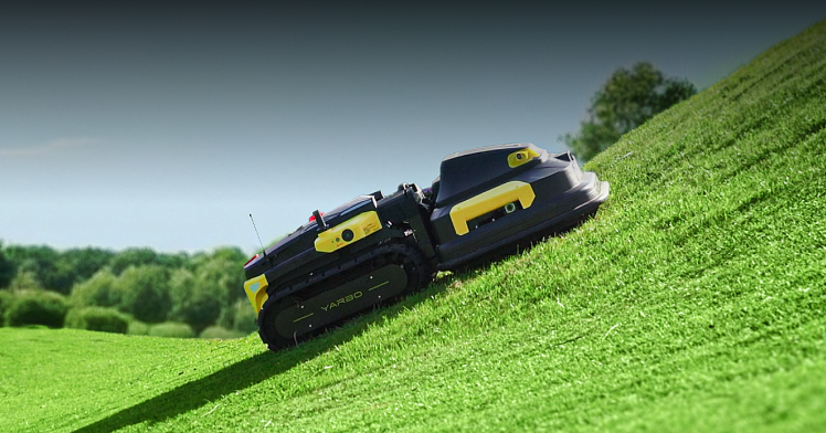 Black and yellow VARBO robotic lawn mower on a grassy hill under a clear sky.
