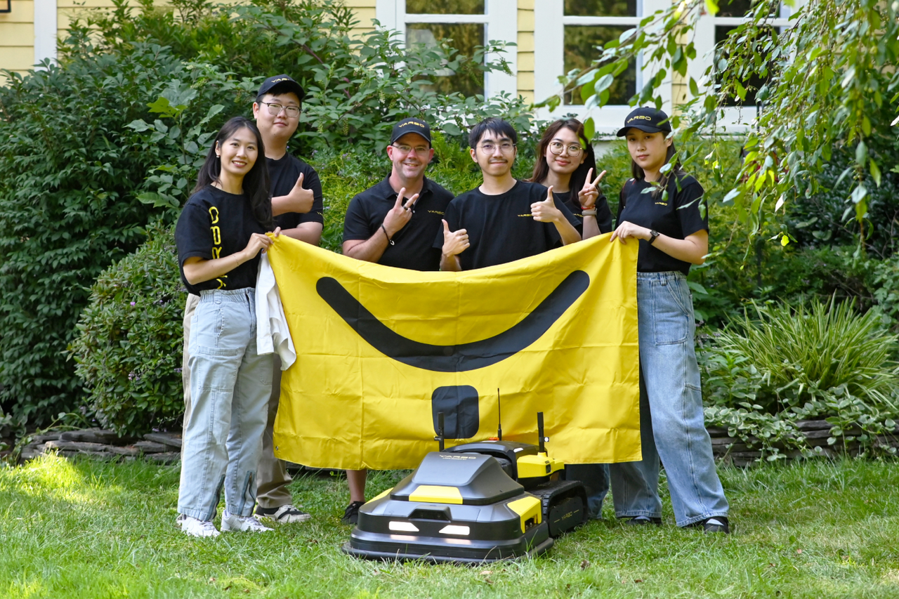 Six people in black shirts and caps pose outdoors with a yellow smiley-face banner, holding thumbs-up and peace signs; two robotic mowers sit in front on grass.