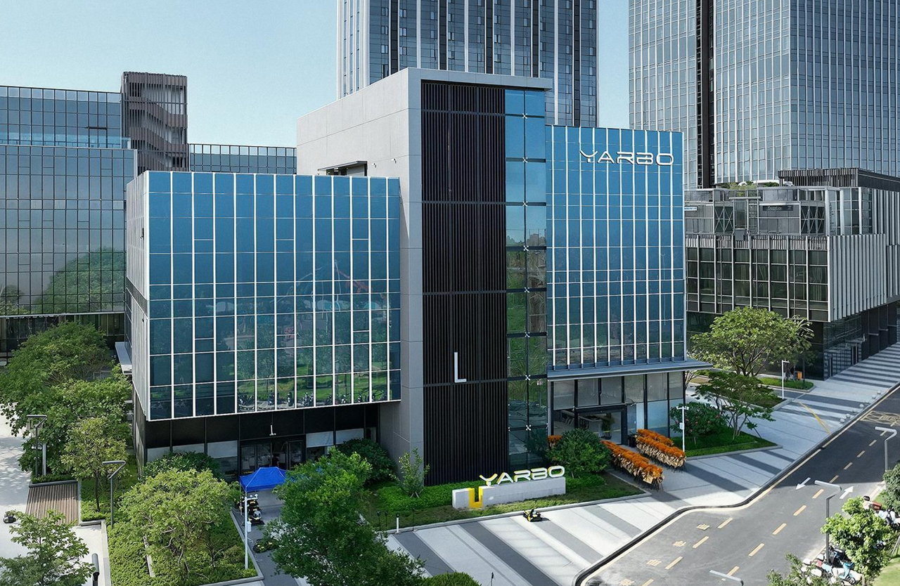 Modern glass YARBO building with blue facade, surrounded by greenery and urban skyscrapers under clear sky.