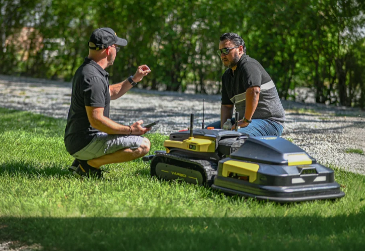 Two men crouch on grass beside yellow and black robotic lawn mowers, discussing features under bright sunlight.