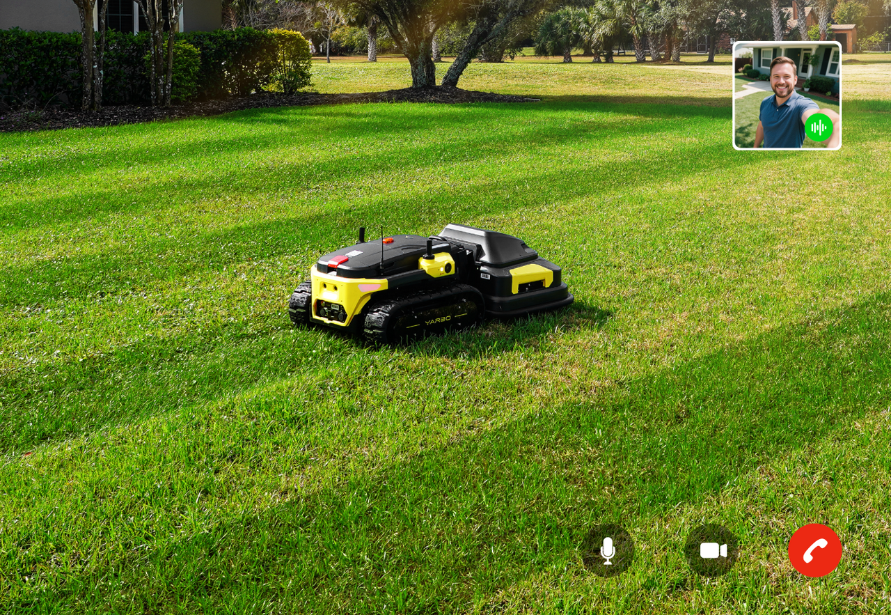 Black and yellow robotic lawn mower on green grass with video call interface overlay showing a smiling man.