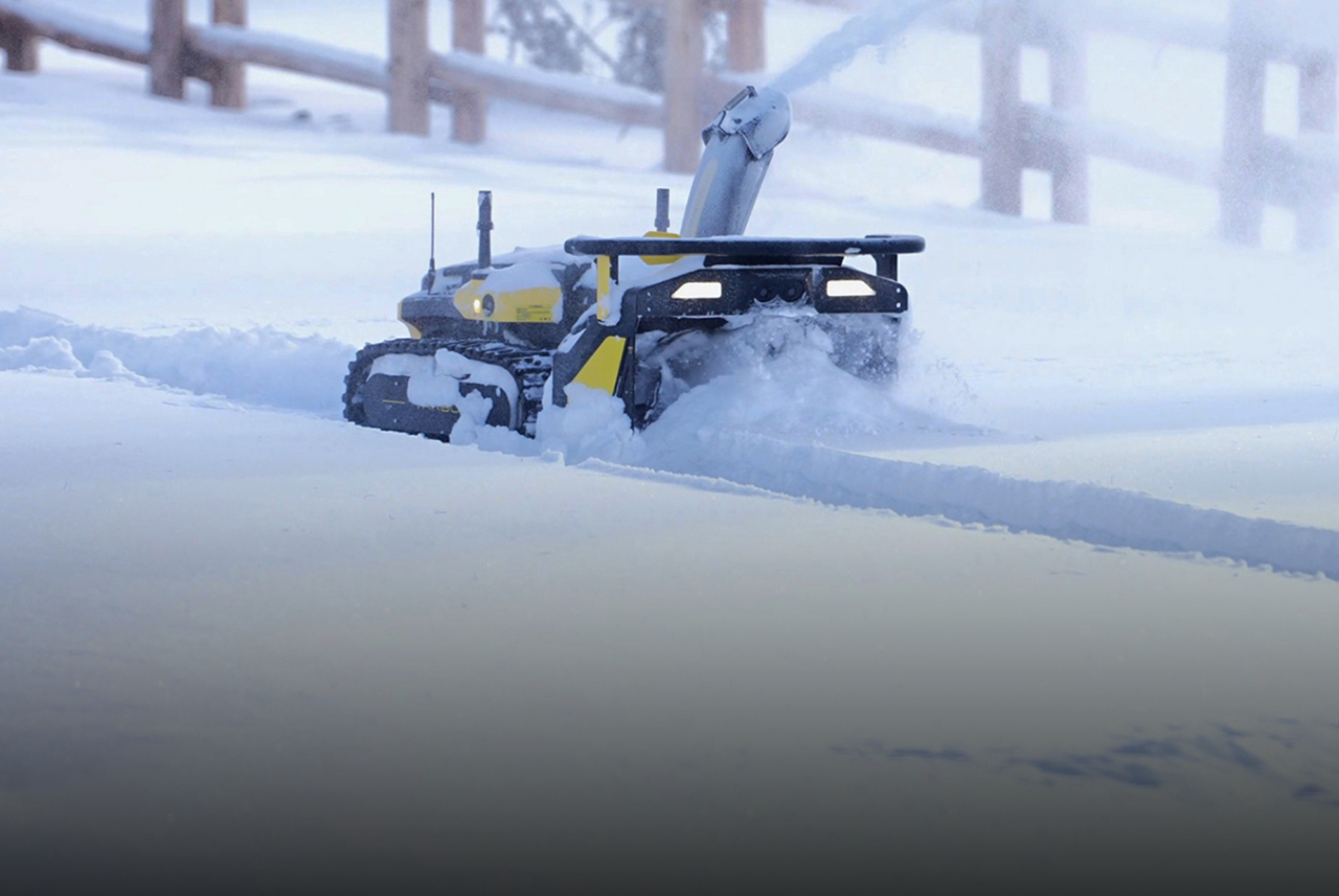 Yellow and black tracked robot plows snow in winter, kicking up snow spray, with wooden fence in background.