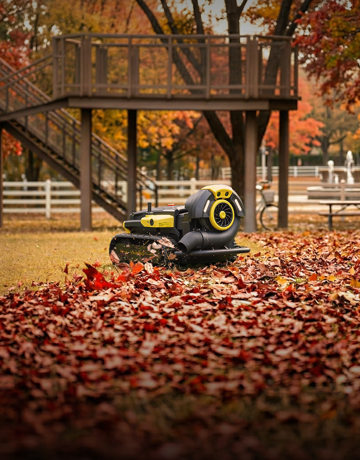 Black and yellow robotic leaf blower on grass covered in red and brown autumn leaves, with wooden deck and trees in background.