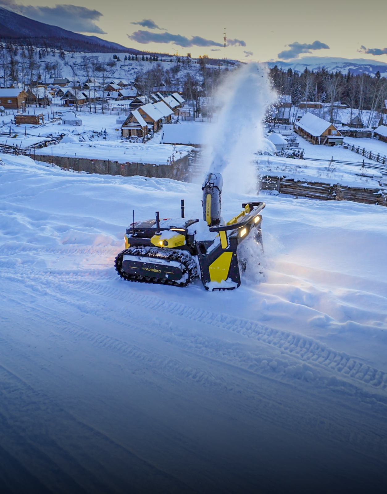 Yellow and black snowblower clearing snow in a snowy village at dusk, with wooden cabins and mountains in the background.
