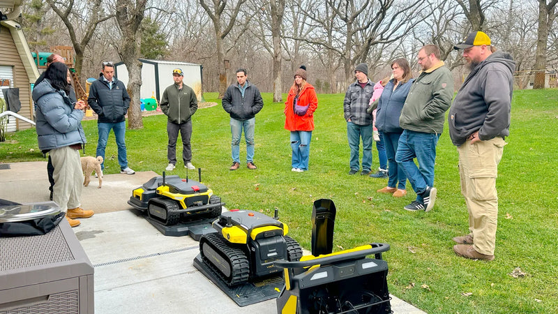 Group observes yellow and black robotic lawn mowers on patio, surrounded by grass and bare trees.