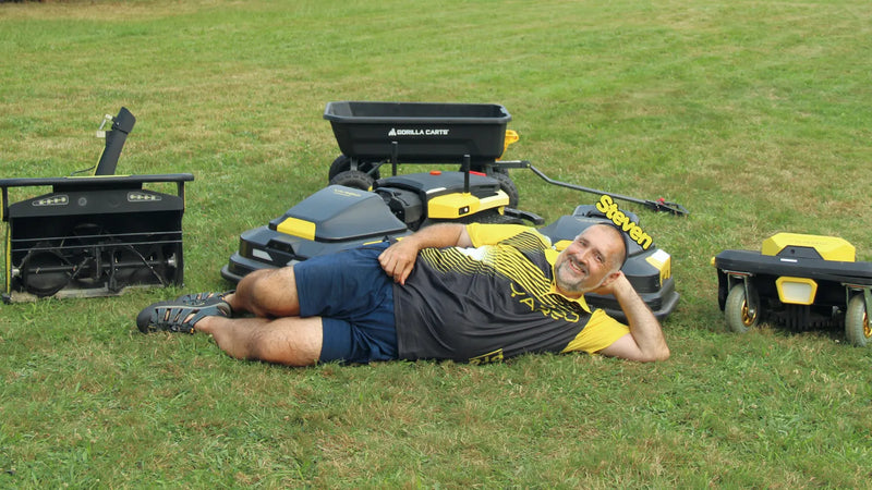 Man in black and yellow shirt labeled "Steven" lies on grass surrounded by yellow and black lawn equipment, including a Gorilla Carts wagon.