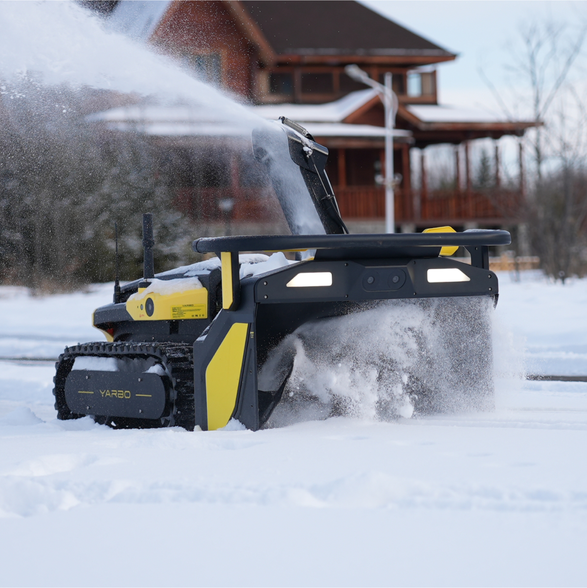 Black and yellow YARBO robotic snow blower clearing snow in front of a wooden house on a snowy day.