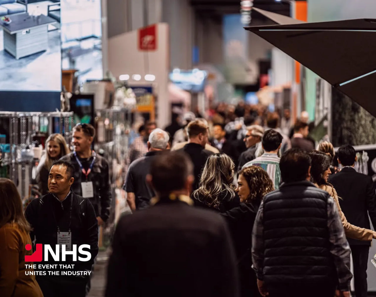 Crowd at NHS trade show, attendees walking between booths, warm lighting, logo in corner.